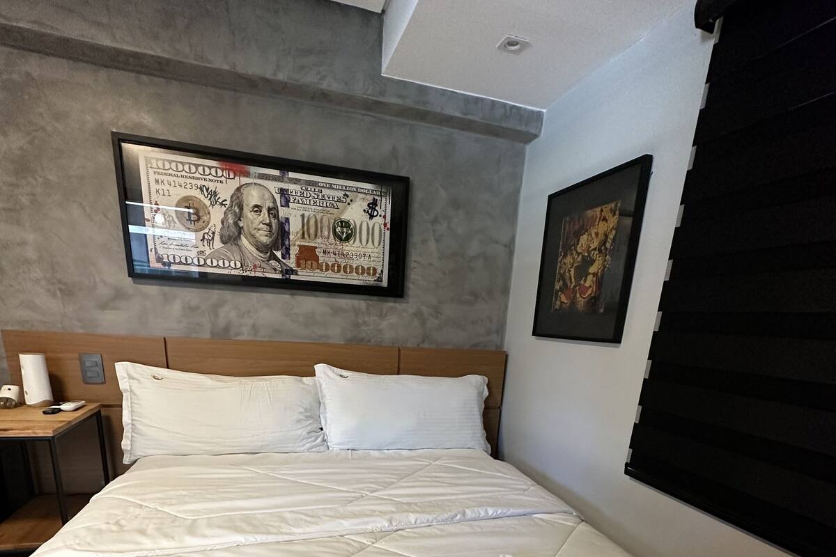A hotel room with a large bed, a framed dollar bill artwork on the wall, and a window with blinds.