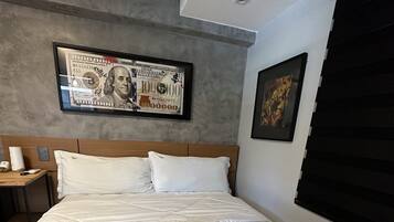 Una habitación de hotel con una cama grande, un cuadro con un billete de dólar enmarcado en la pared y una ventana con persianas.