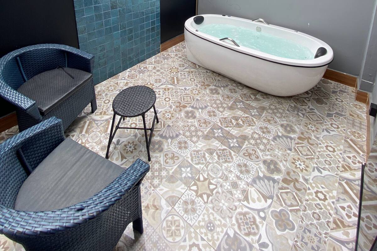 A bathroom with a bathtub, two wicker chairs, a small stool, and patterned floor tiles.