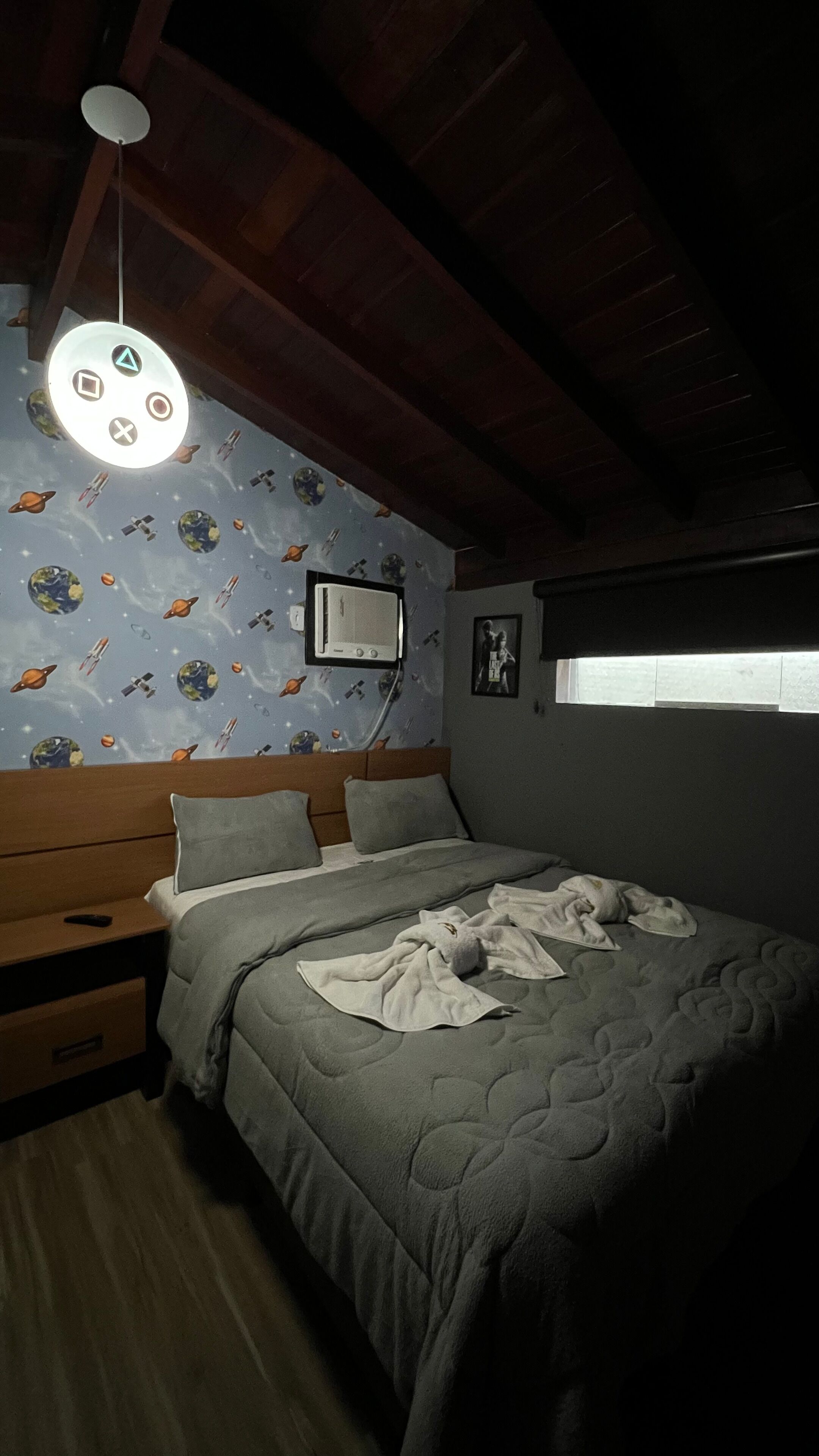 Quarto com teto de madeira, cama com colcha cinza, mesa de cabeceira, aquecedor fixado na parede e papel de parede com tema espacial.