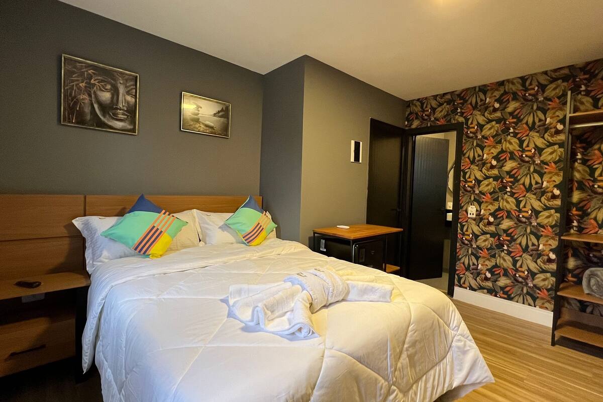 A bedroom with a bed, a wooden nightstand, a small table, a mirror, and a floral wallpaper.