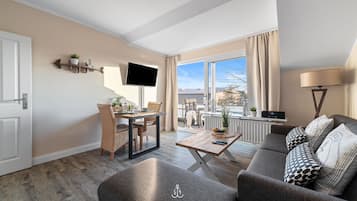 Una sala de estar moderna con un sofá, una mesa de comedor y un televisor. Se ve el exterior a través de grandes ventanales.