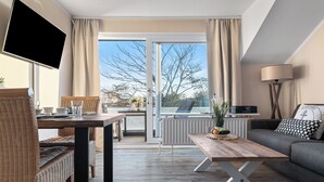 Un soggiorno moderno con divano, tavolino in legno, tavolo da pranzo con sedie, televisione a parete e vista sull'esterno tramite una porta in vetro.