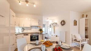 A modern kitchen with white cabinetry, a dining table set for breakfast, and a cozy seating area with a chair and a small table. - Suutje Bude - for 4 persons, terrace, quiet location (Büsum)
