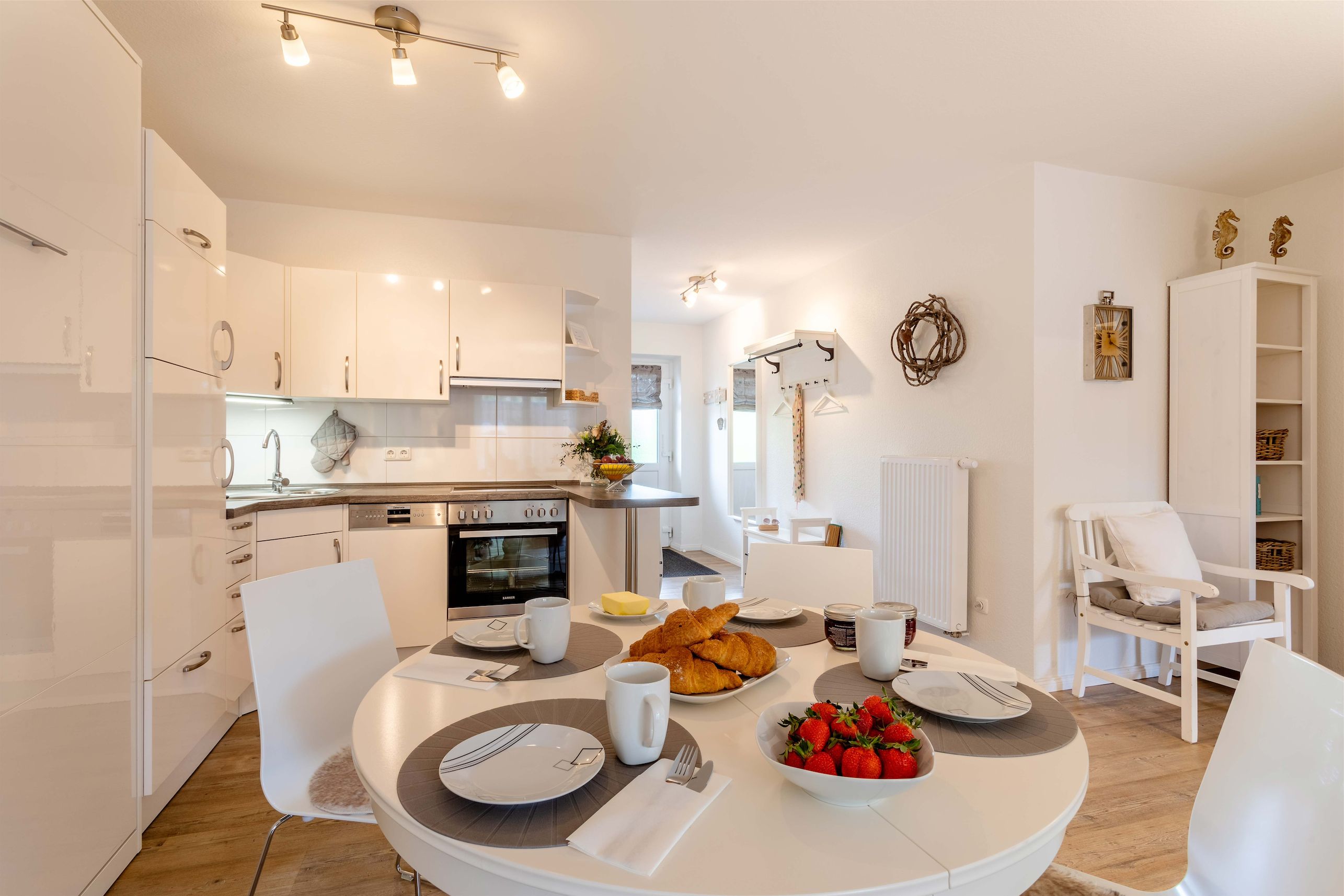 Una cocina moderna con armarios blancos, una mesa de comedor lista para el desayuno y un rincón acogedor con una silla y una mesita.