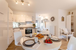 Une cuisine moderne avec des meubles blancs, une table à manger dressée pour le petit-déjeuner et un coin salon confortable avec un fauteuil et une petite table.