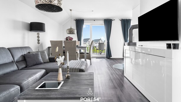 En moderne stue med en grå sofa, et sofabord med en tablet, en hængende lampe og udsigt til det fri gennem store vinduer.