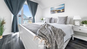 Et moderne soveværelse med en stor seng, hvide sengeborde, et natbord med lampe og udsigt til bygninger gennem vinduet.