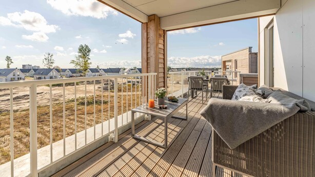 A balcony with a wicker sofa, a small table with a drink, and a view of houses and trees. - Gode Tied - Fireplace, Infrared Sauna, Bathtub, Shower, Balcony, Parking Space (Olpenitz)