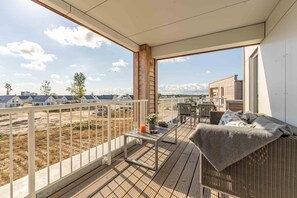 A balcony with a wicker sofa, a small table with a drink, and a view of houses and trees.
