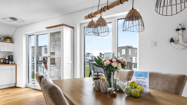 A modern kitchen with white cabinets, a wooden dining table, and a view of neighboring buildings. - Strandmuschel 65-Balcony, Infrared Sauna, Shower, Fireplace, EV Charging Station (Olpenitz)