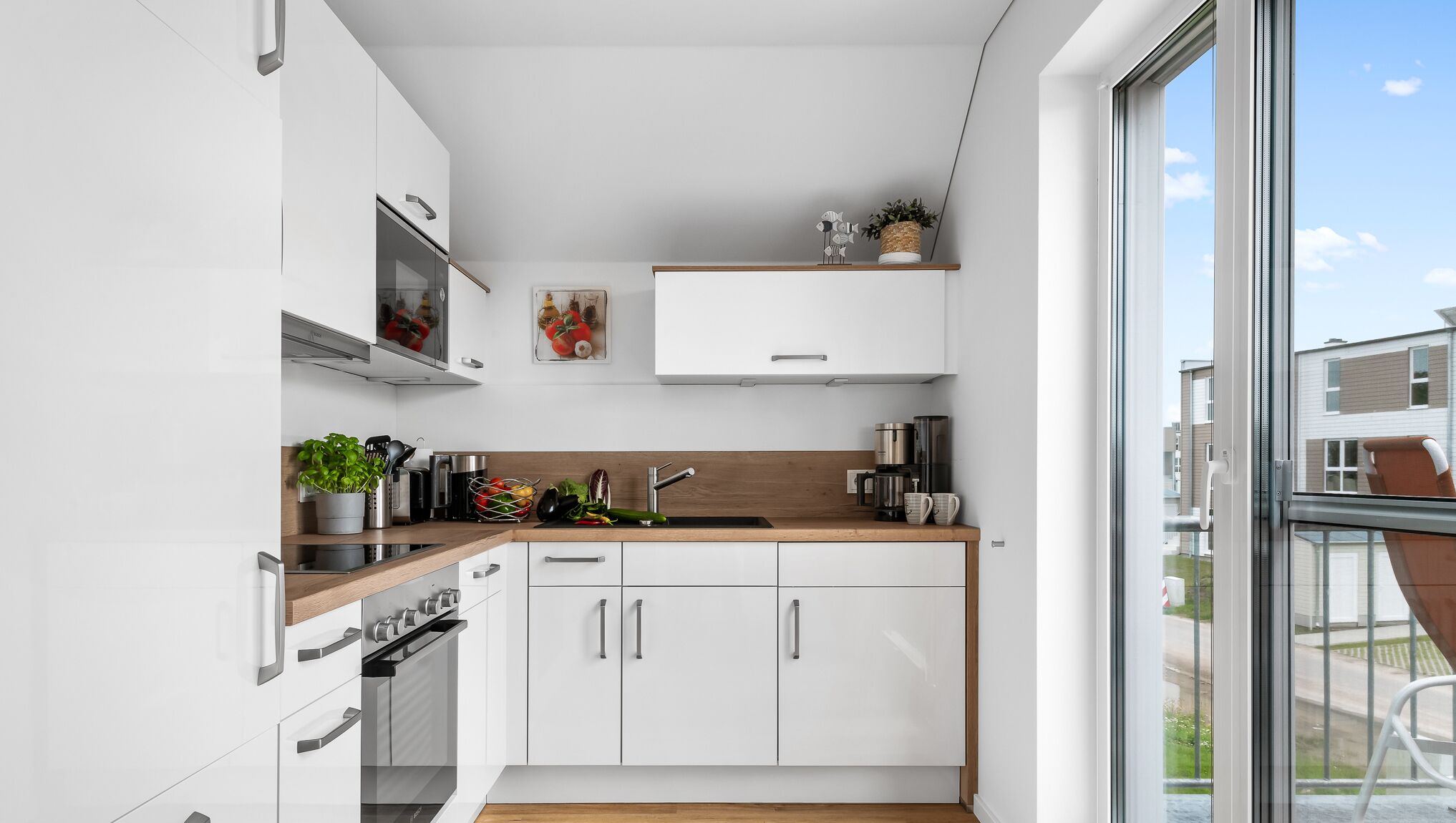 A modern kitchen with white cabinets, a wooden countertop, and a large window offering a view of the neighborhood.