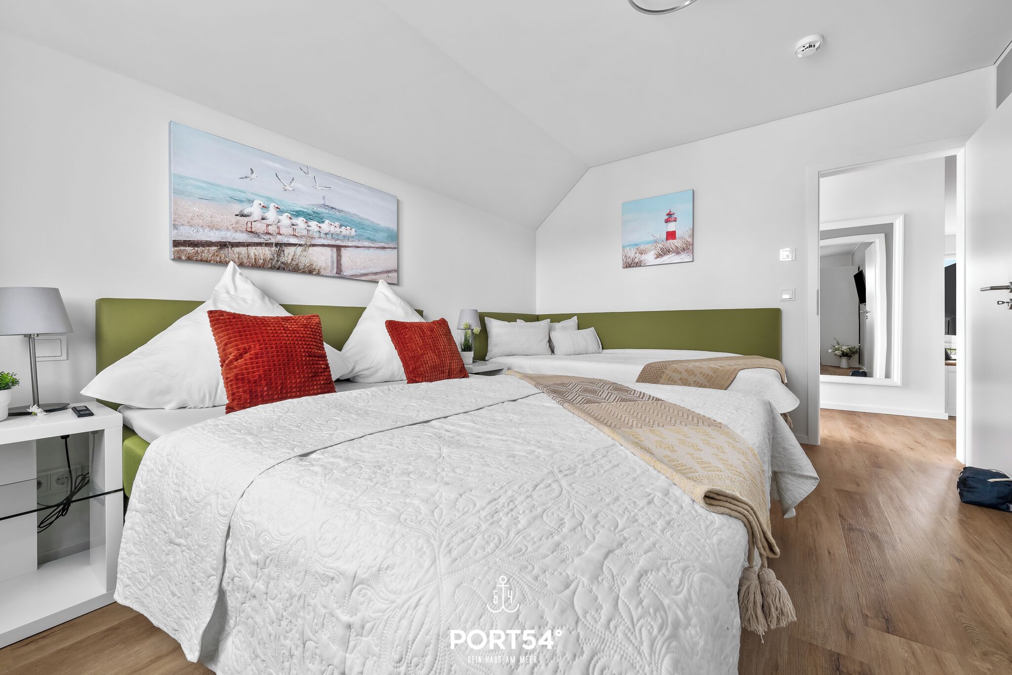 Ein modernes Hotelzimmer mit einem großen Bett, zwei Nachttischen und einem Beistelltisch. An den Wänden hängen zwei Bilder, auf dem Beistelltisch steht eine Lampe und eine Tür führt in einen anderen Raum.