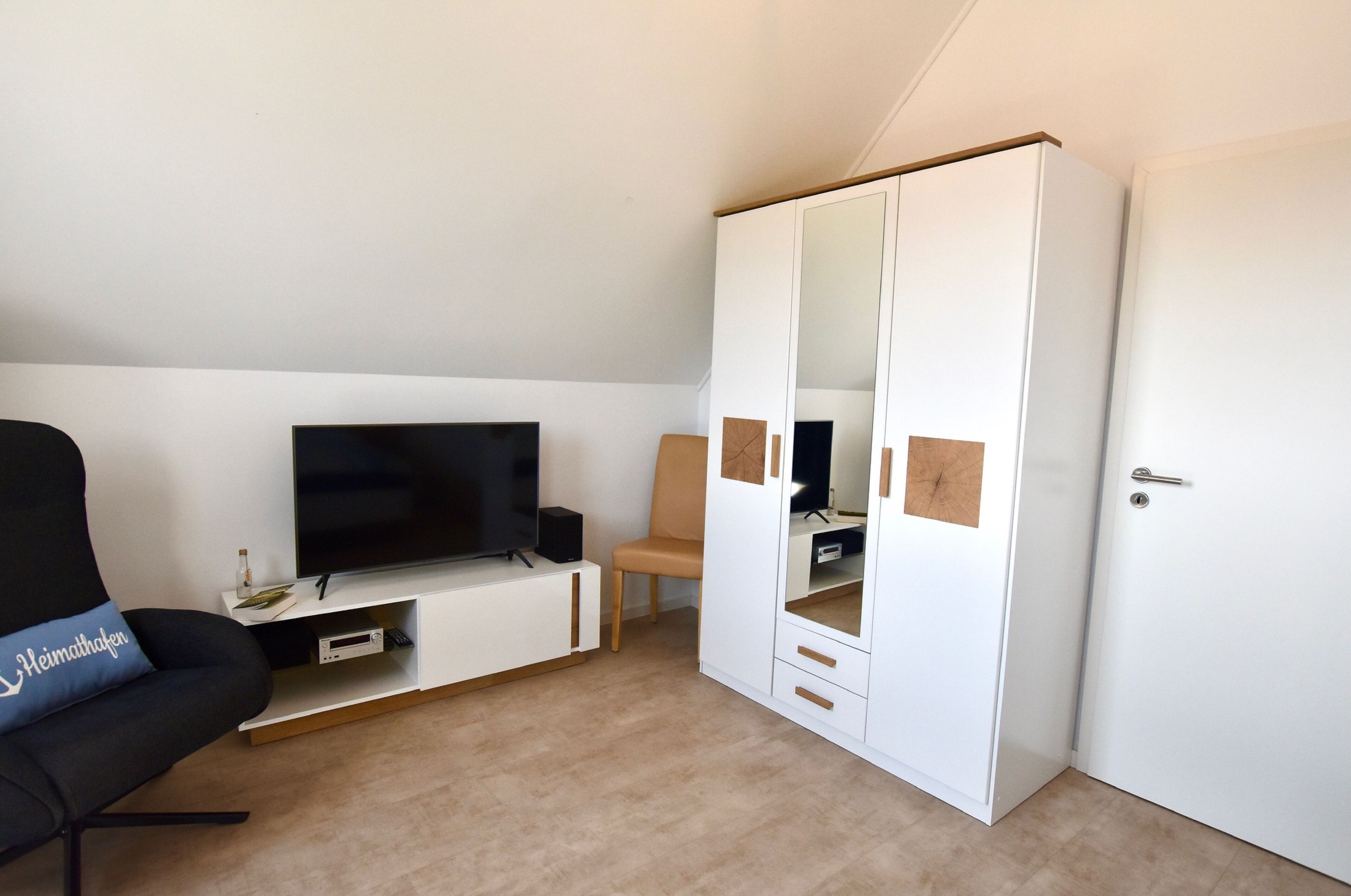 A room with a TV on a white stand, a black chair with a pillow, a wooden chair, and a white wardrobe with a mirror.