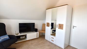 A room with a TV on a white stand, a black chair with a pillow, a wooden chair, and a white wardrobe with a mirror.