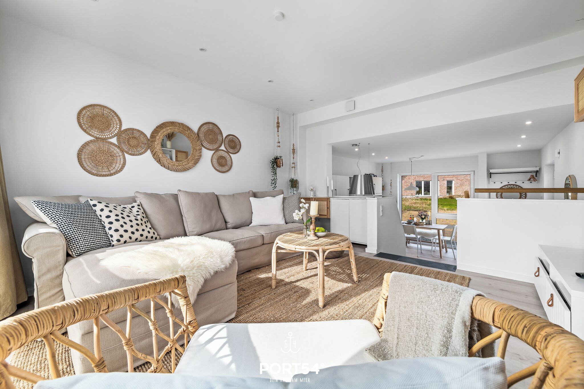 Een moderne woonkamer met een bank, rieten stoelen en een ronde salontafel. Achtergrond toont een keuken.