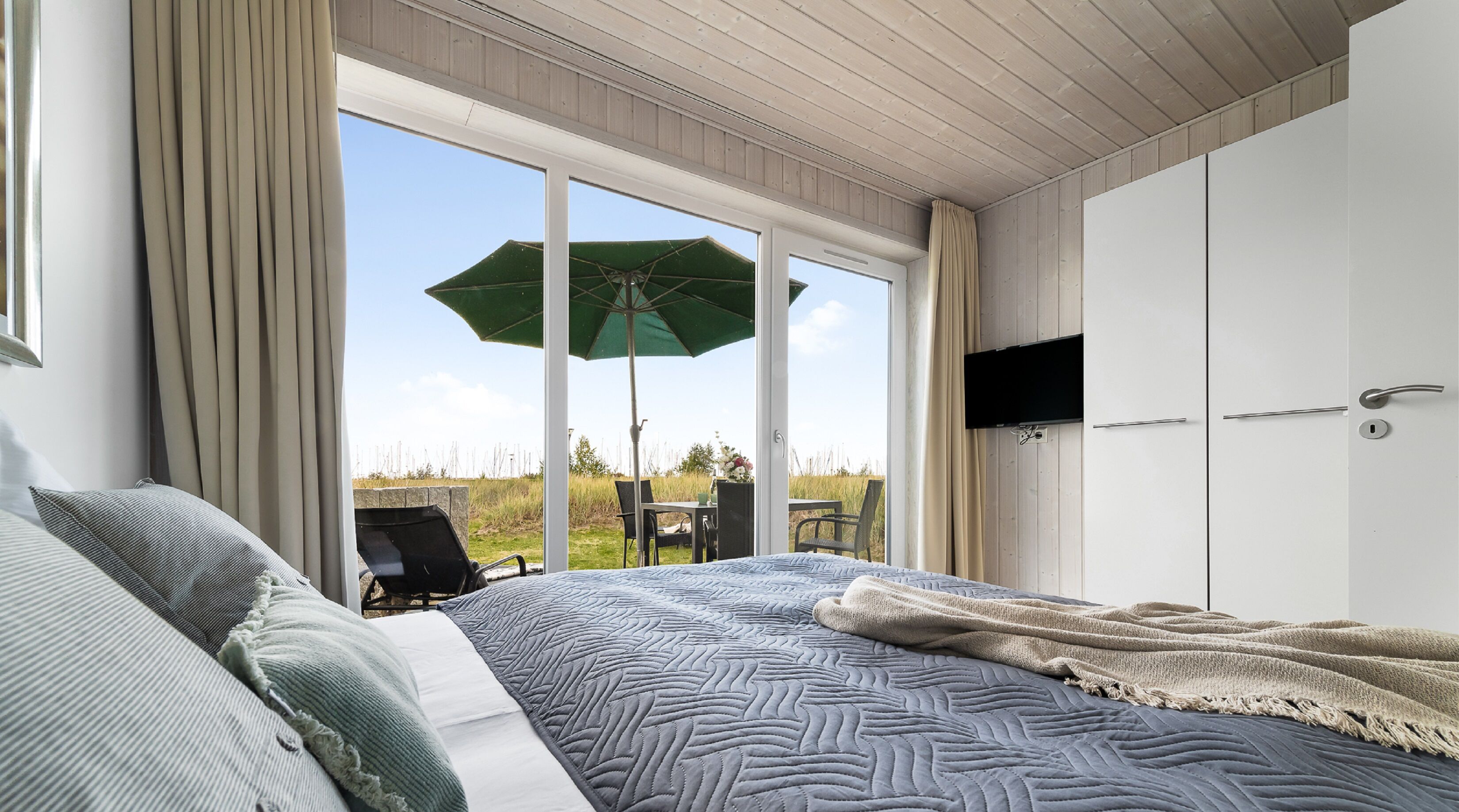 Ein Schlafzimmer mit einem großen Bett, einem an der Wand montierten Fernseher und einer Glastür, die zu einem Außenbereich mit Tisch und Stühlen führt.