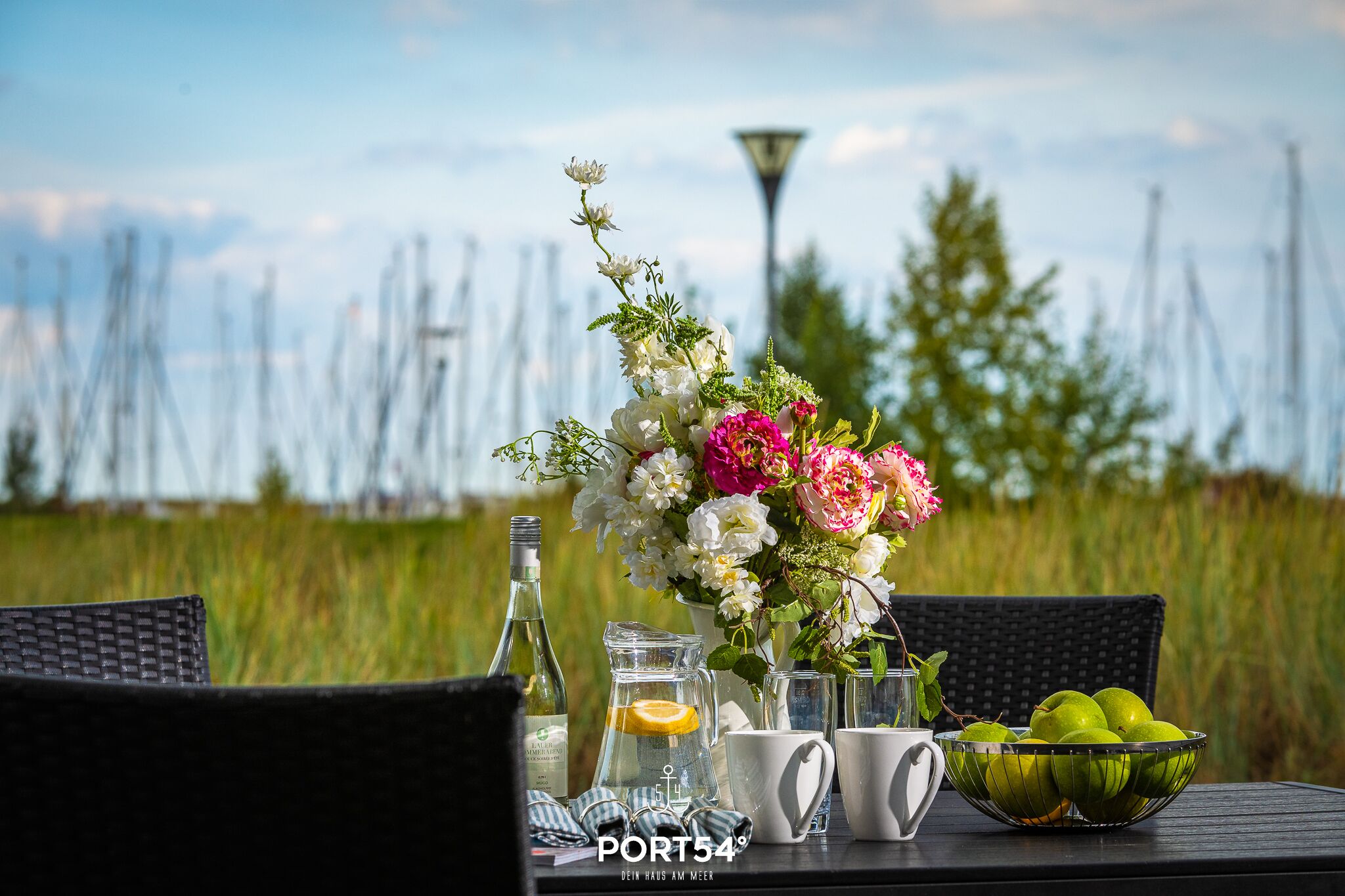 A table set outdoors with a floral arrangement, a pitcher, glasses, a bowl of apples, and a bottle.