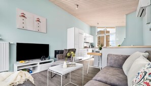 A modern living room with a flat-screen TV, a sofa, a coffee table, and a kitchen area visible through an open door.