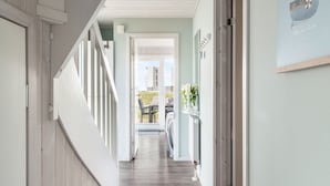 A hallway with a wooden floor, white walls, and a staircase on the left. There is a door on the right leading to a room with a bed and a window.