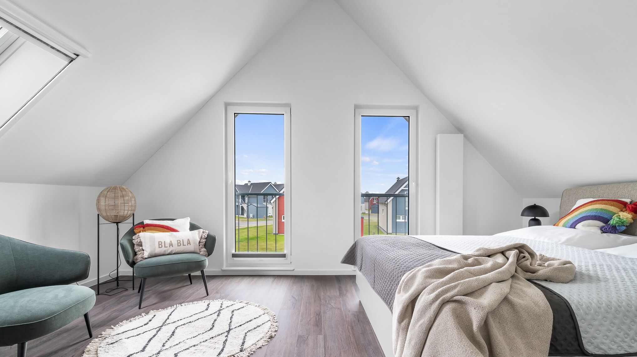 Une chambre avec un plafond en pente, deux fenêtres, un lit, une chaise, une lampe et une vue sur les maisons à l’extérieur.
