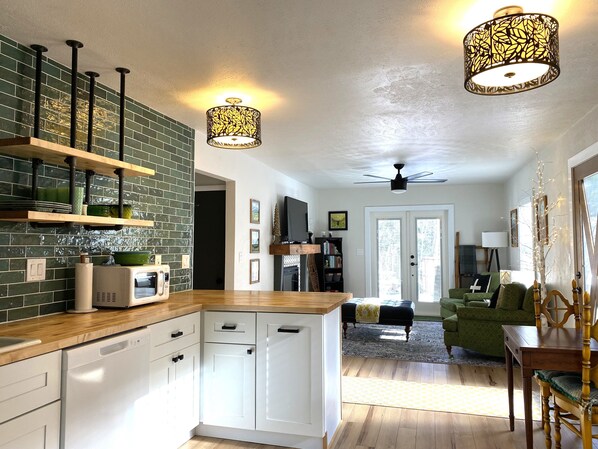 Una cocina moderna con armarios blancos, encimera de madera y salpicadero verde. Hay una zona de comedor con un sofá verde y una mesa de madera.