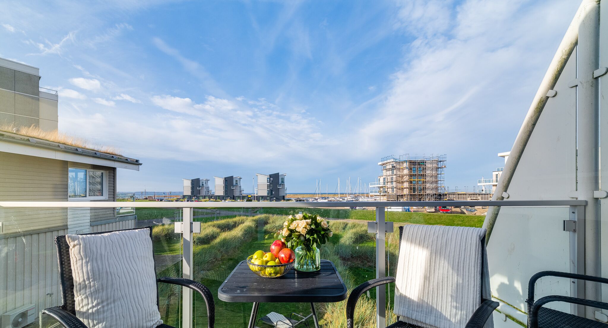 A balcony with a table set with fruits and flowers, overlooking a marina with boats and buildings.