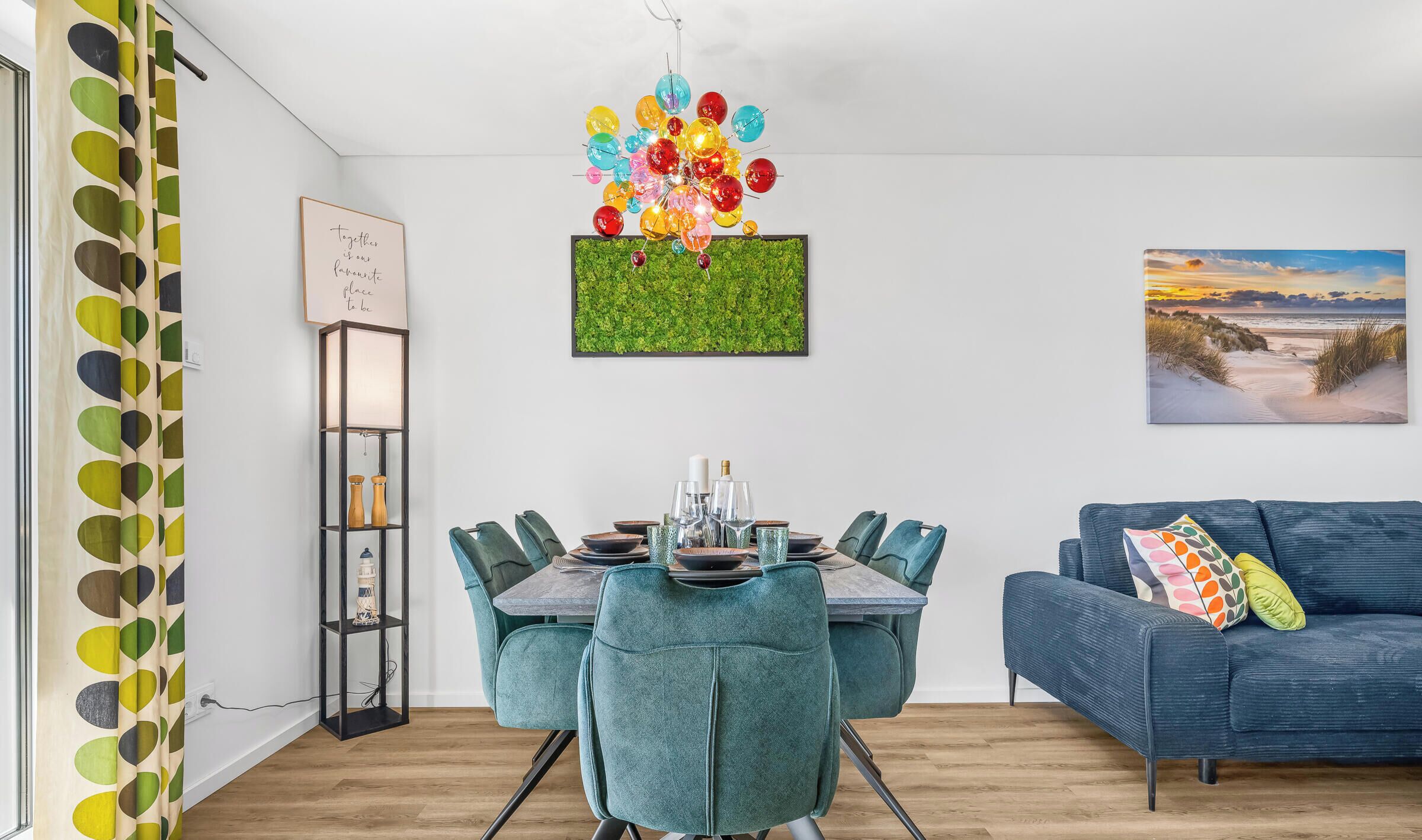 Un coin repas avec une table dressée pour six personnes, un canapé bleu, un luminaire fixé au mur et un tableau encadré.