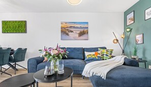 A modern living room with a blue sofa, a dining area with chairs, a coffee table, and a wall-mounted picture of a beach scene. - Tüddelbüddel - Infrared Sauna, Bathtub, Fireplace, Terrace, Grill, Parking (Olpenitz)