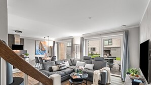 A modern living room with a grey sofa, a coffee table, and a dining area with a table and chairs. There is a staircase leading to another level, and a large window with curtains. - Ferienhaus Hannes Büsum (Büsum)