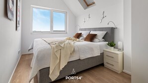 A modern bedroom with a grey bed, white bedding, a bedside table with a plant, and a window with a view of the sky. - Fjordblick– first row on the water, sauna, whirlpool, shower, fireplace, balcony (Olpenitz)
