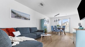 Ein modernes Wohnzimmer mit grauem Sofa, einem Esstisch mit Stühlen, einem kleinen weißen Couchtisch und einer Küchenzeile mit blauem Schrank.