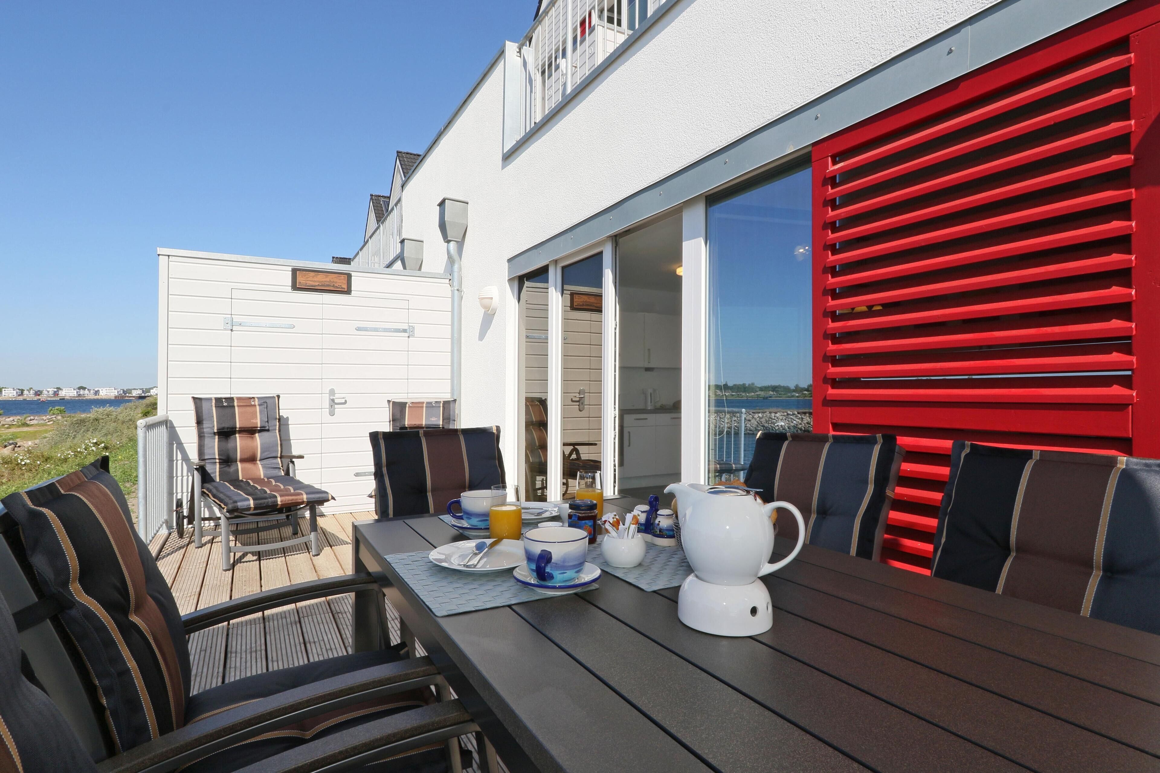 An outdoor dining area with a table set for breakfast, including cups, a teapot, and a pitcher. There are chairs with striped cushions, and a red awning is visible.