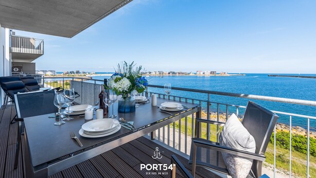 Mesa de comedor dispuesta con platos, vasos y cubiertos en una terraza al aire libre con vista al mar y edificios a lo lejos.