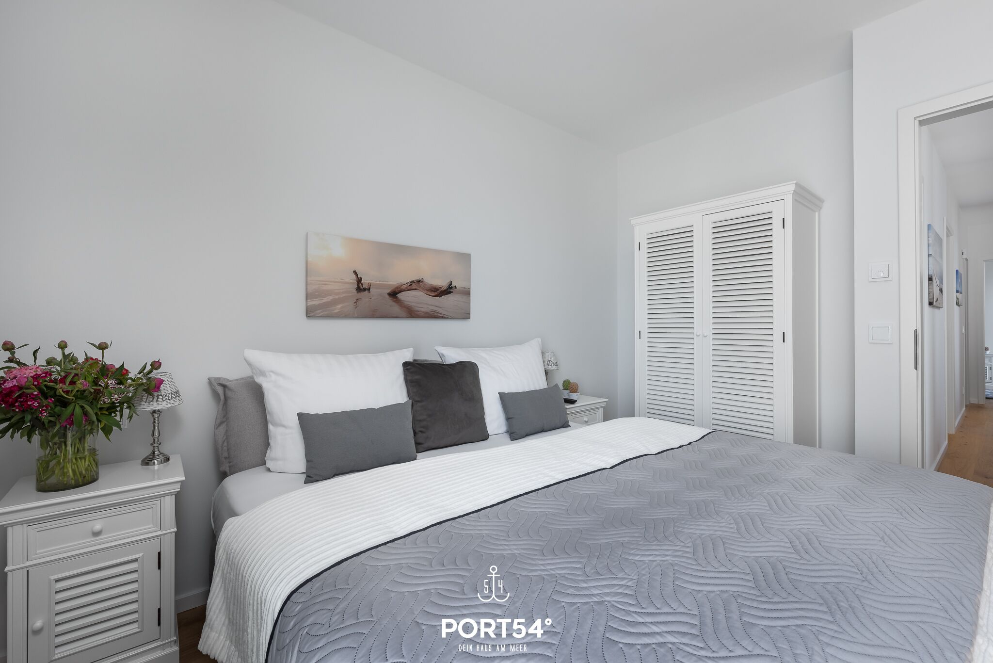 Ein Schlafzimmer mit einem Bett, weißen Nachttischen, einem Blumenstrauß, einem Bild an der Wand und einem Kleiderschrank.