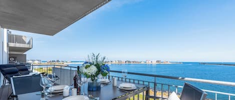 A dining table set with plates, glasses, and cutlery on an outdoor deck with a view of the sea and buildings in the distance.