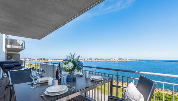 A dining table set with plates, glasses, and cutlery on an outdoor deck with a view of the sea and buildings in the distance.