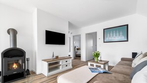 Uma sala de estar moderna com uma lareira a lenha Justus, uma TV de tela plana, um rack branco, um sofá e um quadro de uma catedral.