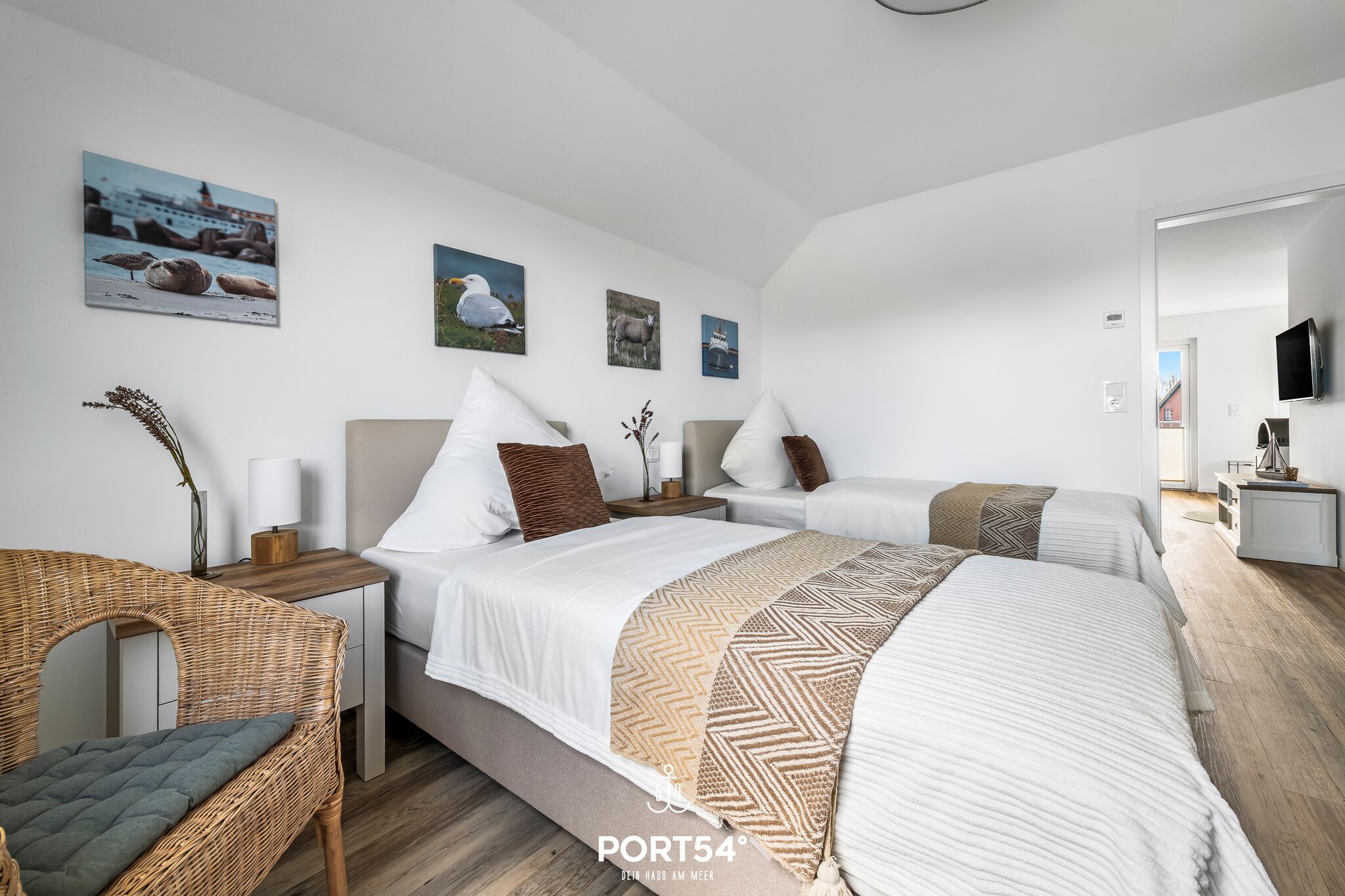 Ein Hotelzimmer mit zwei Betten, einem Korbstuhl, einem Nachttisch mit Lampe und Wandbildern von Möwen und Schafen.
