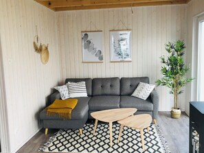 Un salon chaleureux avec un canapé d’angle gris, un plaid jaune, deux tables basses en bois, un tapis à motifs noir et blanc et une grande plante verte.