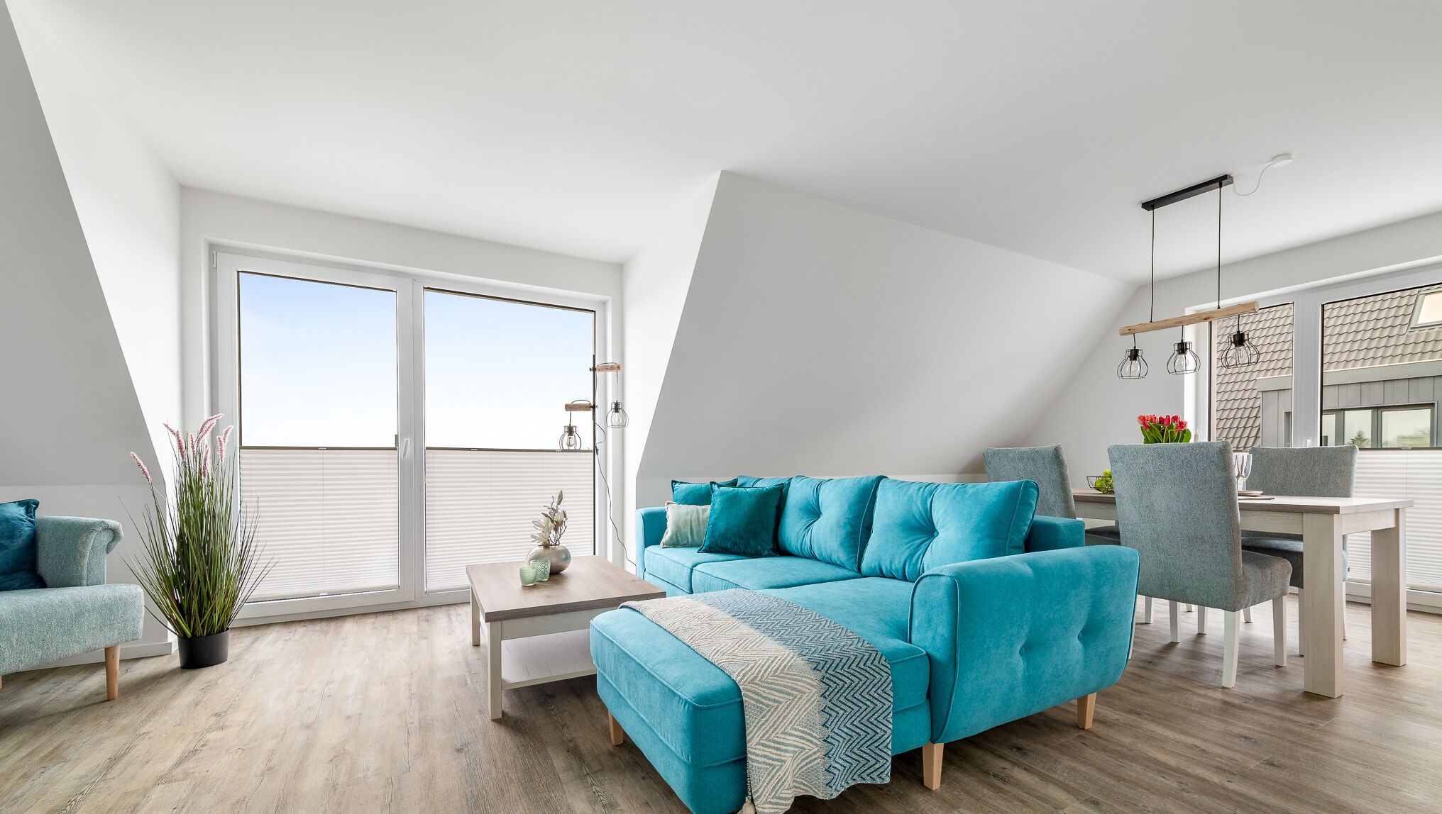 Ein modernes Wohnzimmer mit einem türkisfarbenen Sofa, einem Essbereich und einem großen Fenster mit Blick auf den Himmel.