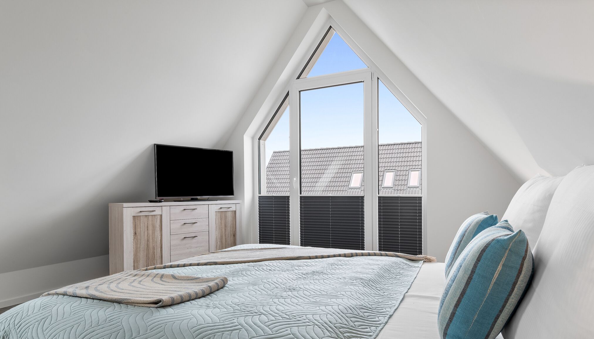 A bedroom with a sloped ceiling, a TV on a wooden dresser, a bed with a quilt, and a view of buildings through a large window.