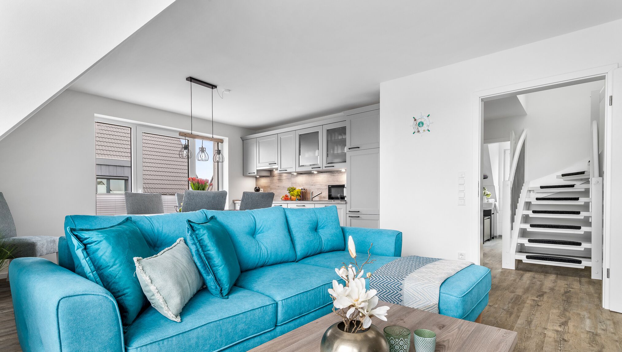 A modern living room with a blue sofa, a wooden coffee table, and a view of the kitchen and staircase.