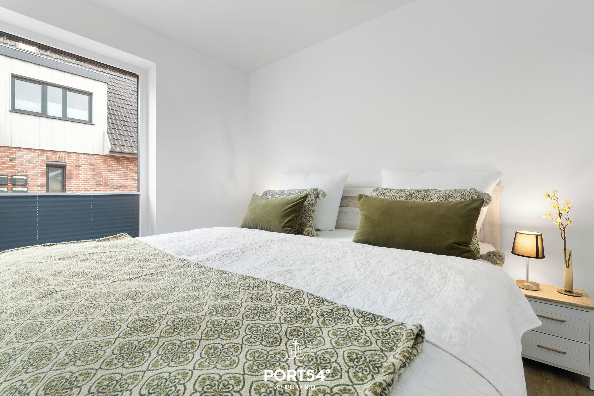 A modern bedroom with a large bed, a nightstand, and a window with a view of a brick building.