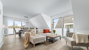 Un vaste espace de vie comprenant un canapé, une table basse et des fauteuils, de grandes fenêtres et une cuisine à l’arrière-plan.