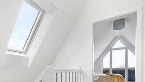 Une pièce aux hauts plafonds, dont un est en pente, avec une lucarne et un lit recouvert d’une couverture jaune.