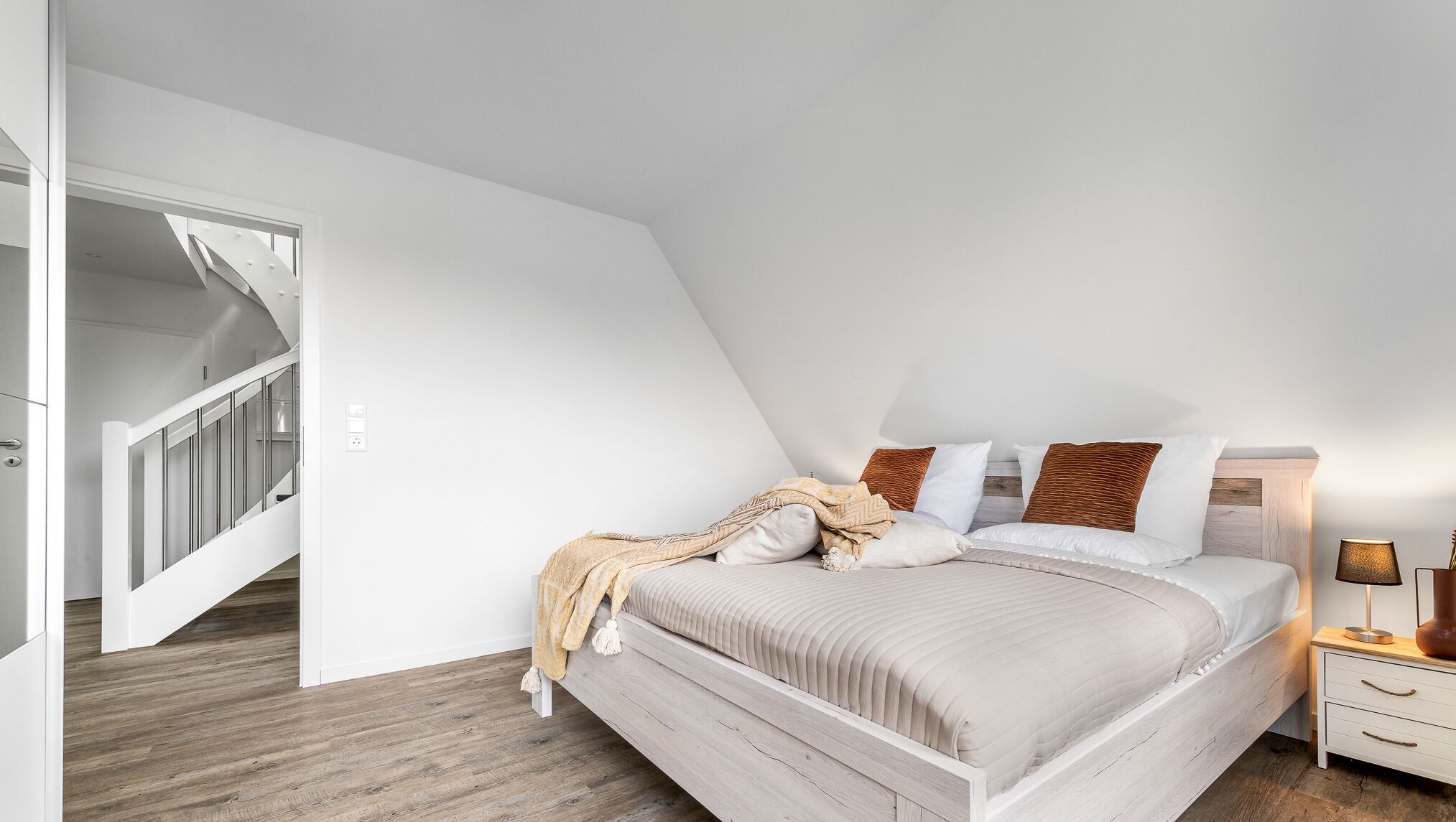 Quarto com teto inclinado, cama com roupa de cama branca e almofadas marrons, luminária de cabeceira e piso de madeira.