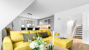 A modern living room with a yellow sofa, a dining area with a table and chairs, a kitchen with grey cabinets, and a staircase leading to another level. - Coast 8 - 2 bedrooms, shower room, balcony, stylishly furnished (Westerdeichstrich)