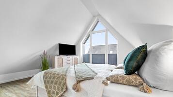 Ein Schlafzimmer mit schräg gedämmter Decke, einem Bett mit gemusterten Betttextilien, einem Fernseher auf einer Holzkommode und einem großen Fenster mit Blick ins Freie.