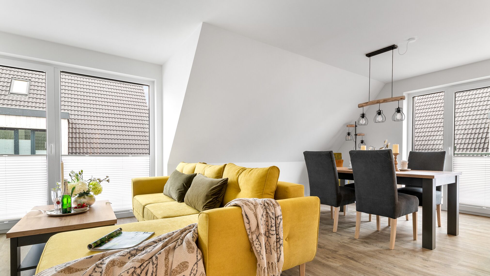 A modern living room with a yellow sofa, a dining table with chairs, and large windows with blinds.